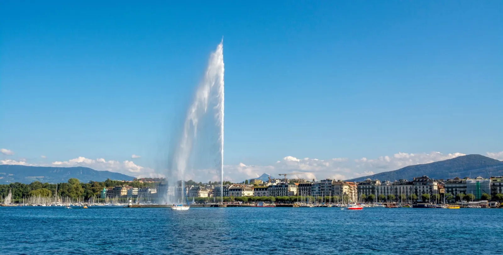 Panoramic view of the Geneva Jet d'Eau on Lake Geneva, with buildings behind and mountains on the horizon