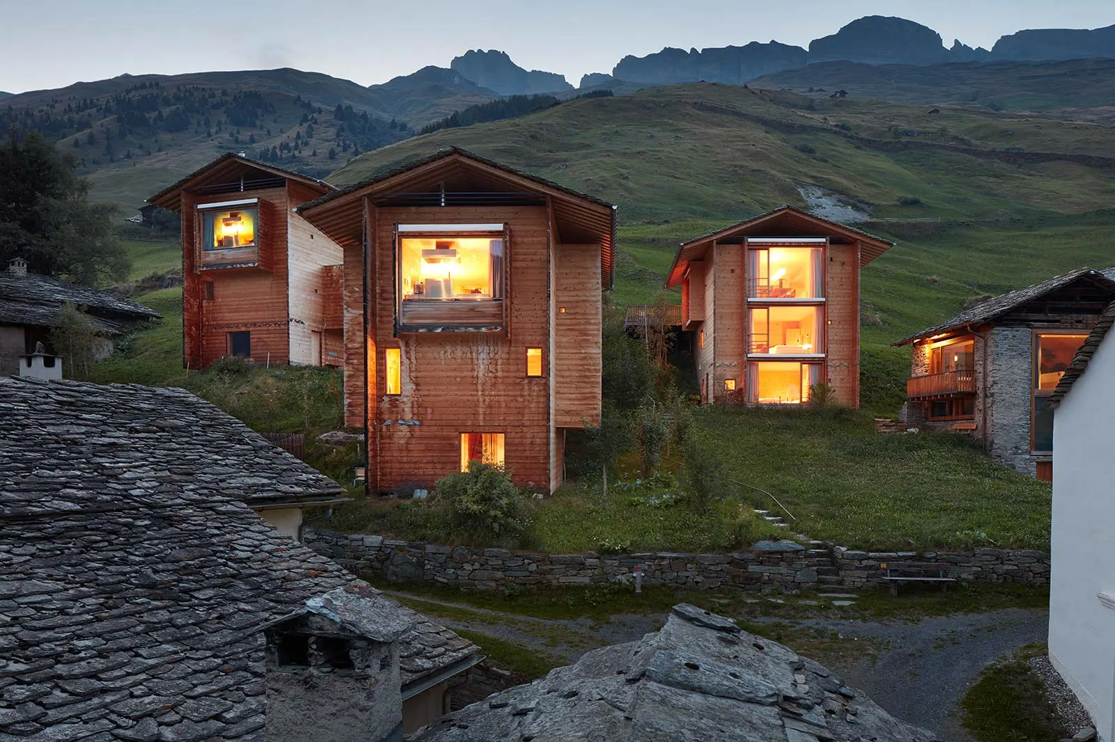 The three wooden houses designed by Peter Zumthor in the hamlet of Leis, near Vals, lit up at nightfall in the mountain landscape