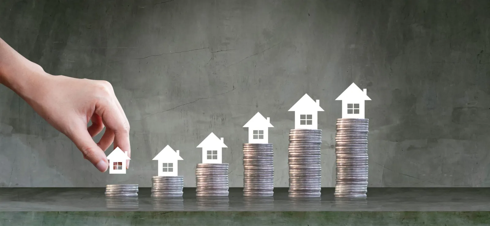 Stacks of coins growing taller, topped with miniature houses, a hand placing the first house