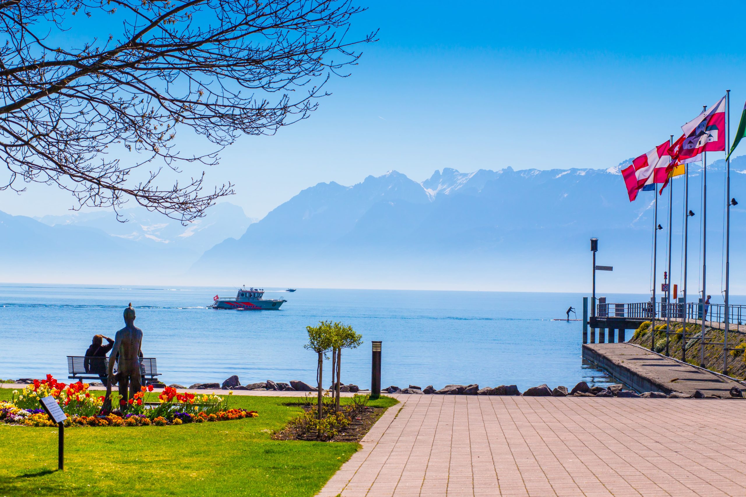 Promenade fleurie au bord du lac Léman à Saint-Sulpice avec bateau et montagnes en arrière-plan.
